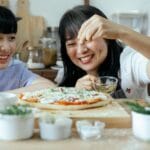 Mama i córka posypują przyprawami domową pizzę w przytulnej kuchni